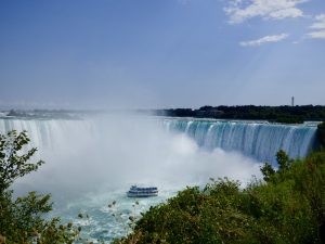 Niagarafälle unter blauem Himmel. Ein winzig anmutendes Ausflugsboot ist direkt vor den tobenden Wasserfällen.