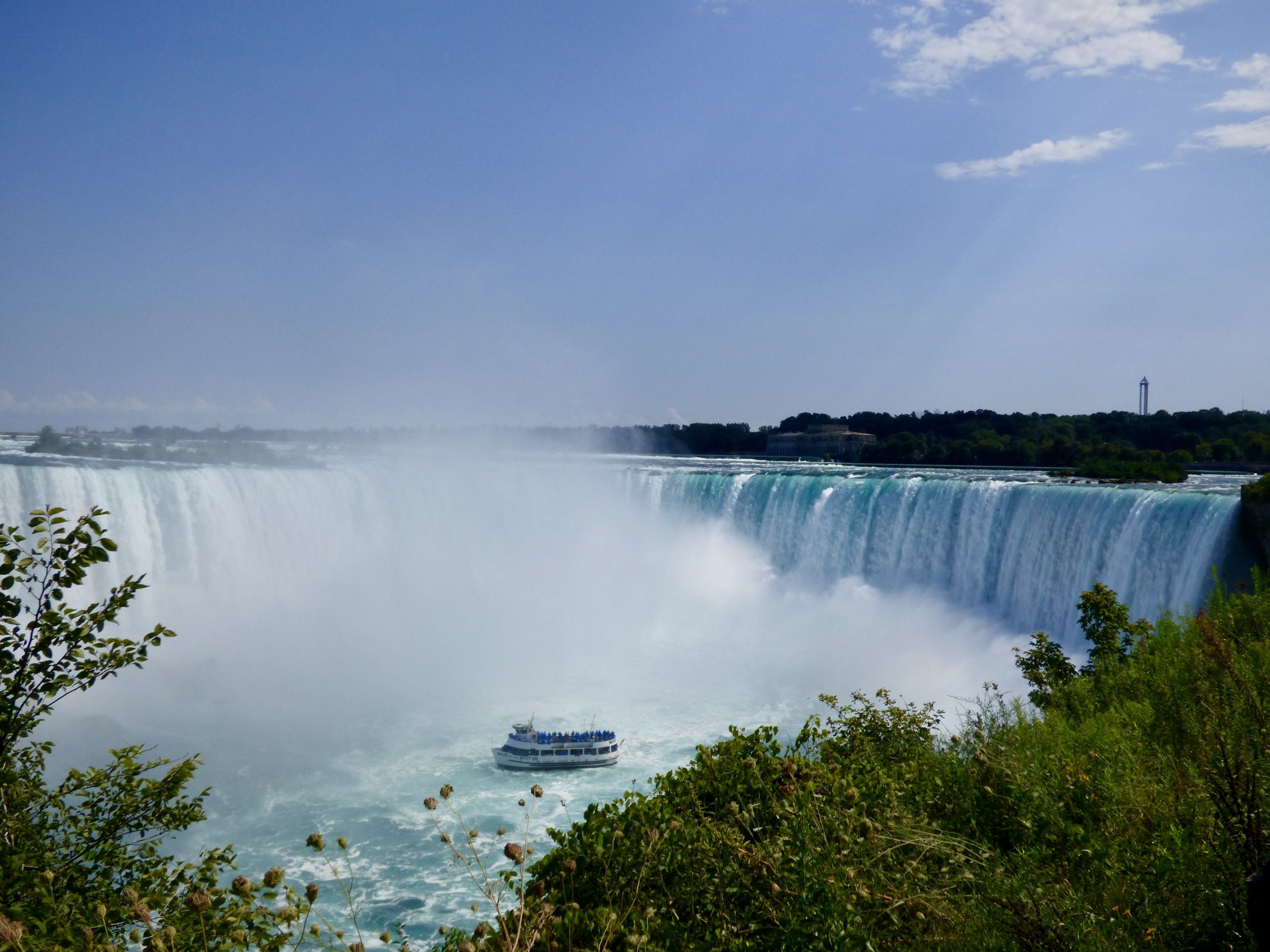 Niagarafälle unter blauem Himmel. Ein winzig anmutendes Ausflugsboot ist direkt vor den tobenden Wasserfällen.