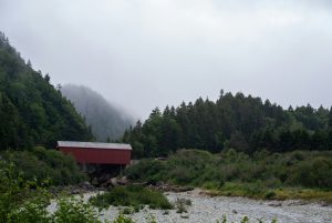Rote, überdachte Brücke vor bewaldeten Hügeln unter grauem Himmel